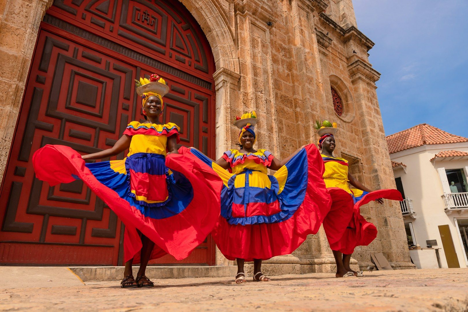 Three women in vibrant traditional dresses pose in front of a grand, ornate door.
